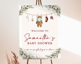 May include: A baby shower welcome sign on a black metal easel. The sign has a cream background with the text "WELCOME TO Samantha's BABY SHOWER" in burgundy script. Decorative elements include a garland and baby clothes.