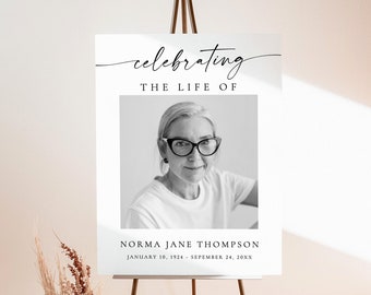 May include: A black and white photo of a woman with glasses is on a white poster with the text "celebrating the life of Norma Jane Thompson January 10, 1924 - September 24, 20XX". The poster is on an easel.