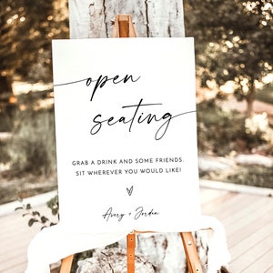 May include: White sign with black calligraphy that reads "open seating" and "Grab a drink and some friends. Sit wherever you would like!" with a heart and the names "Avery + Jorden" below. The sign is on a wooden easel.