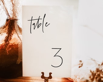 Op de afbeelding: Witte kaart met zwarte kalligrafie met de tekst "tafel 3" op een houten oppervlak met een gouden metalen standaard.