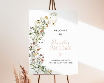 May include: A white welcome sign with a floral design featuring a variety of flowers and greenery. The text reads "Welcome to Deniella's Baby Shower" with the date "September 14th, 20XX" and the phrase "We're so glad you're here!" below. The sign is on a gold easel.