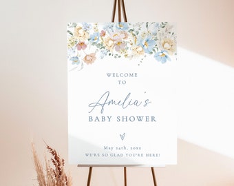 May include: A welcome sign for a baby shower with a floral design in shades of blue, yellow, and pink. The sign reads "Welcome to Amelia's Baby Shower" and includes the date "May 24th, 20xx". The sign also says "We're so glad you're here!" and has a small heart at the bottom. The sign is on a wooden easel.