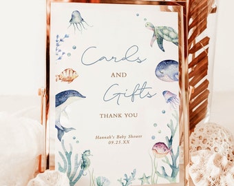 May include: A framed sign with a gold-colored metal frame. The sign reads "Cards and Gifts" with "Thank You" below. Watercolor illustrations of sea creatures, including a dolphin, jellyfish, and seahorse, decorate the sign. The sign is for Hannah's Baby Shower.