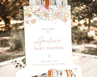 May include: A white welcome sign with floral illustrations at the top. The text reads "Welcome to Amelia's Baby Shower" with a pink heart and the date "May 24th, 20xx". The sign is on a wooden easel.