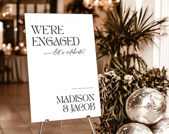 May include: A white 24x36 inch foam board sign on a metal tripod stand. The text reads WE'RE ENGAGED, let's celebrate!, and MADISON & JACOB in black serif and script fonts. The sign is displayed in a room with plants and disco balls.