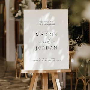 May include: A white foam board sign on a wooden easel, with the text "WELCOME TO THE WEDDING OF MADDIE and JORDAN" and the date "OCTOBER 12, 20XX". The sign is 18x24 inches.