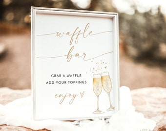 May include: A framed sign with the words "waffle bar" in gold script. Below, it reads "Grab a waffle, add your toppings, enjoy." Two champagne flutes are illustrated on the right. The sign is on a stone surface with a white cloth.