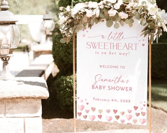 May include: A baby shower welcome sign with a gold metal frame. The sign reads "a little SWEETHEART is on her way!" and "Welcome to Samantha's BABY SHOWER" with a heart design. The sign is decorated with flowers.