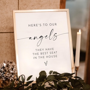 May include: A framed sign with the text "HERE'S TO OUR angels THEY HAVE THE BEST SEAT IN THE HOUSE" in black script. A white candle in a gold holder and greenery are in the foreground. The sign has a gold frame.