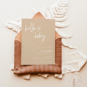 Op de afbeelding: Een beige babyborrel uitnodiging met witte kalligrafie die "hello baby" zegt. De uitnodiging bevat de datum, tijd en locatie van het evenement. De uitnodiging is versierd met een witte varen en een bruine envelop.