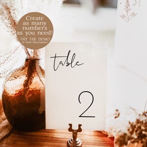 May include: A white card with the word "table" and the number "2" printed in black. The card is on a gold metal stand and is sitting on a wooden table.