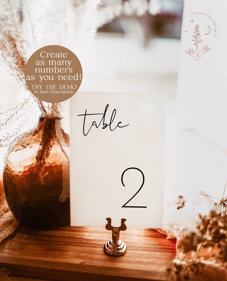 May include: A white card with the word "table" and the number "2" printed in black. The card is on a gold metal stand and is sitting on a wooden table.
