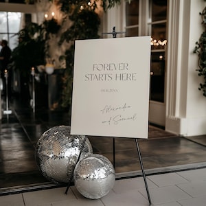 May include: A rectangular off-white wedding sign on a black metal easel featuring the black text FOREVER STARTS HERE, the date 08.16.20XX, and the names Alexandra and Samuel. Two silver mirrored disco balls sit on the floor at the base of the easel.