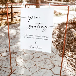May include: A white sign with black text that reads "open seating" and "Grab a drink and some friends. Sit wherever you would like!" with the names "Avery + Jordan" at the bottom. The sign is displayed on a copper stand.