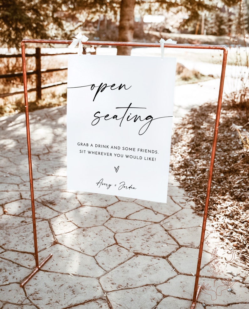 May include: A white sign with black text that reads "open seating" and "Grab a drink and some friends. Sit wherever you would like!" with the names "Avery + Jordan" at the bottom. The sign is displayed on a copper stand.
