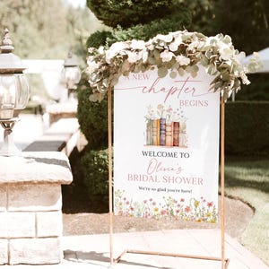 May include: A white foam board sign, 24x36 inches, with a floral and book design, welcomes guests to Olivia's bridal shower. The sign is held by a copper-colored metal frame and decorated with greenery.