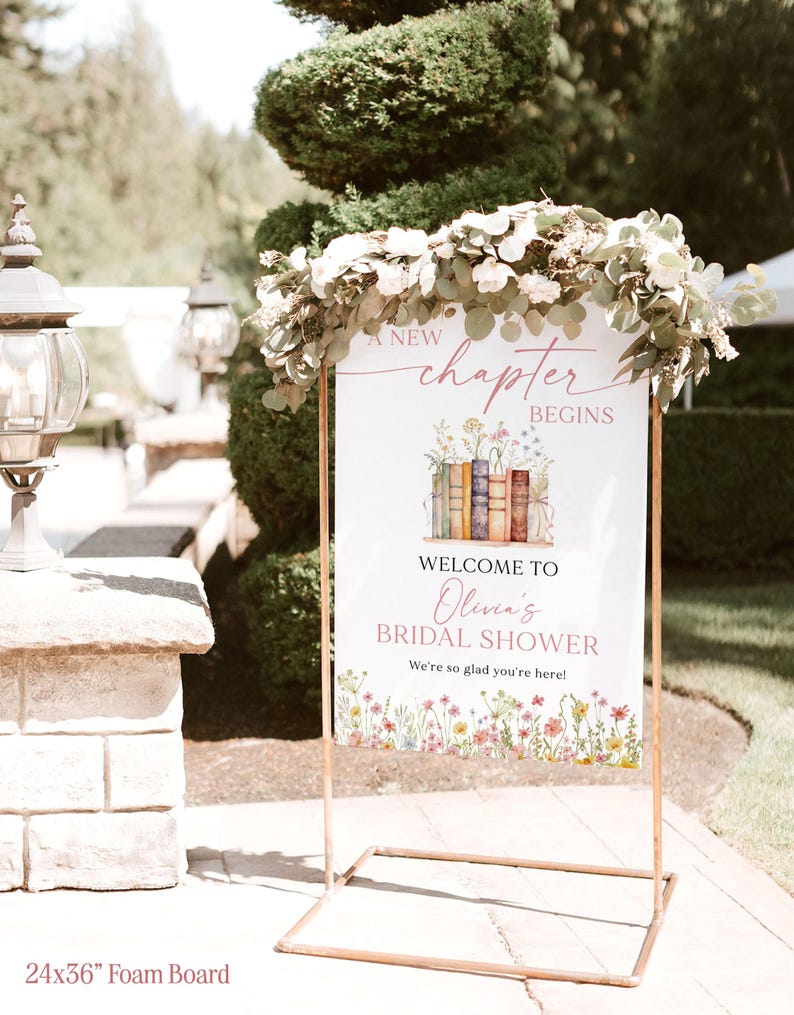 May include: A white foam board sign, 24x36 inches, with a floral and book design, welcomes guests to Olivia's bridal shower. The sign is held by a copper-colored metal frame and decorated with greenery.