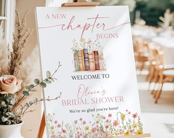 May include: A white 18x24 inch foam board sign on a wooden easel. The text reads A NEW chapter BEGINS, WELCOME TO, Olivia's BRIDAL SHOWER, and We're so glad you're here. It features a watercolor illustration of colorful books and wildflowers.