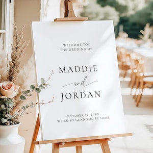 May include: A white 18x24 inch foam board wedding sign on a wooden easel. The text reads: WELCOME TO THE WEDDING OF MADDIE and JORDAN OCTOBER 12, 20XX WE'RE SO GLAD YOU'RE HERE. The sign is displayed next to a white vase with dried flowers.