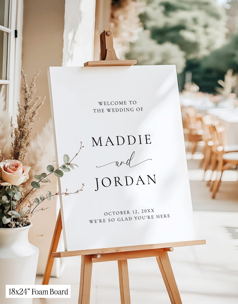 May include: A white 18x24 inch foam board wedding sign on a wooden easel. The text reads: WELCOME TO THE WEDDING OF MADDIE and JORDAN OCTOBER 12, 20XX WE'RE SO GLAD YOU'RE HERE. The sign is displayed next to a white vase with dried flowers.