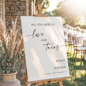 May include: A white rectangular wedding sign on a wooden easel featuring black text that reads ALL YOU NEED IS love AND Tacos AVERY + JORDAN 08.14.20XX | SANTA BARBARA, CA. The sign uses a mix of sans-serif and cursive fonts for the lettering.