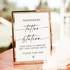 以下が含まれることがあります： 「TEMPORARY tattoo station OUR LOVE IS FOREVER BUT THIS TATTOO ISN'T! PLEASE TAKE ONE」という文字とハートマークが書かれた金色の額縁の看板。木製のテーブルの上に置かれている。