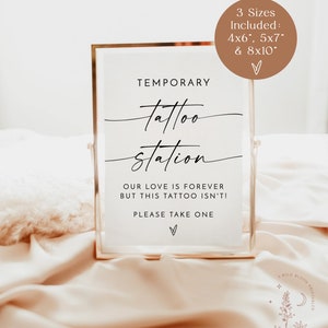 以下が含まれることがあります： 「TEMPORARY tattoo station OUR LOVE IS FOREVER BUT THIS TATTOO ISN'T! PLEASE TAKE ONE」というテキストと、下部に小さなハートマークが描かれた、金色のフレームのサイン。サインは白とピンクのテクスチャのある表面に置かれています。サインは、10 x 15 cm、12.7 x 17.8 cm、20.3 x 25.4 cmの3つのサイズがあります。