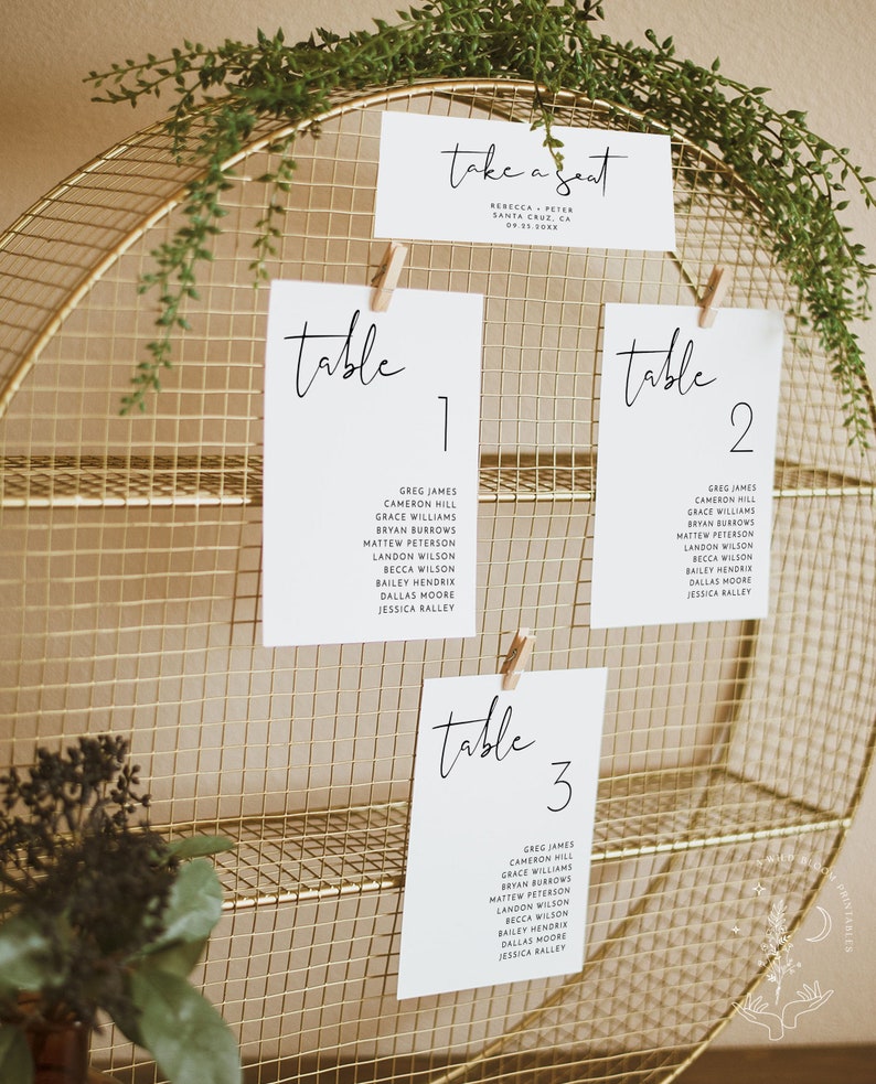 Pu&ograve; includere: Un supporto in filo dorato con tre ripiani che sostengono delle carte bianche con calligrafia nera. Le carte sono etichettate "tavolo 1", "tavolo 2" e "tavolo 3" e elencano i nomi per ogni tavolo. La carta superiore recita "take a seat" con i nomi Rebecca e Peter e la localit&agrave; Santa Cruz, CA.