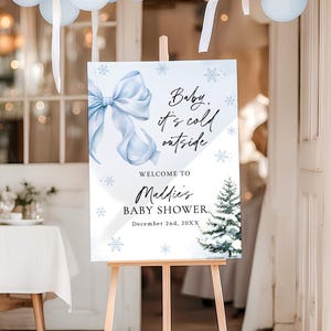 May include: A baby shower sign on a wooden easel. The sign features a watercolor blue bow, snowflakes, and a snowy Christmas tree. Text reads "Baby, it's cold outside" and welcomes guests to "Maddie's Baby Shower" on December 2nd, 20XX.