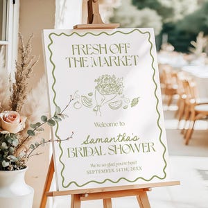 May include: A white foam board sign on a wooden easel with the text "FRESH OFF THE MARKET" and "Samantha's BRIDAL SHOWER" in green. The sign has a green wavy border and a floral illustration. The sign is 18x24 inches.