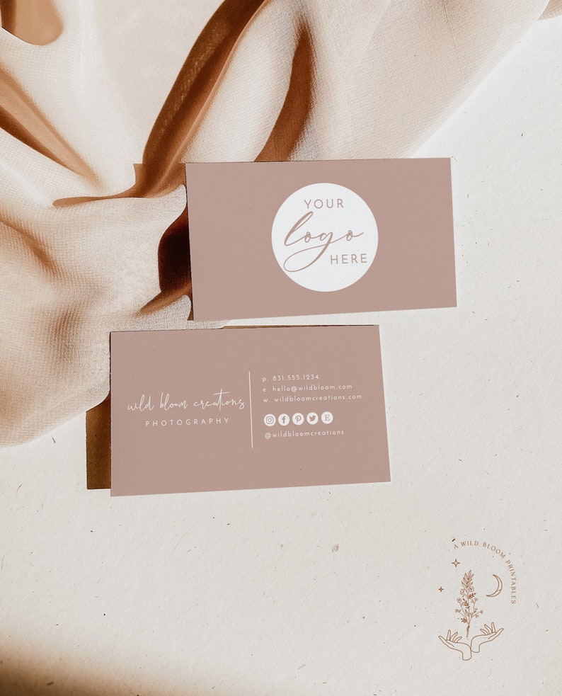 May include: A business card with the text "wild bloom creations PHOTOGRAPHY" and contact information, including a phone number, email address, website, and social media links. The card is light brown with white text. The background is a light brown fabric.