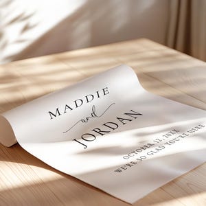 May include: A white poster paper featuring black printed text that reads MADDIE and JORDAN, followed by the date OCTOBER 12, 20XX and the phrase WE'RE SO GLAD YOU'RE HERE. The paper is partially rolled at one end on a light wood surface.