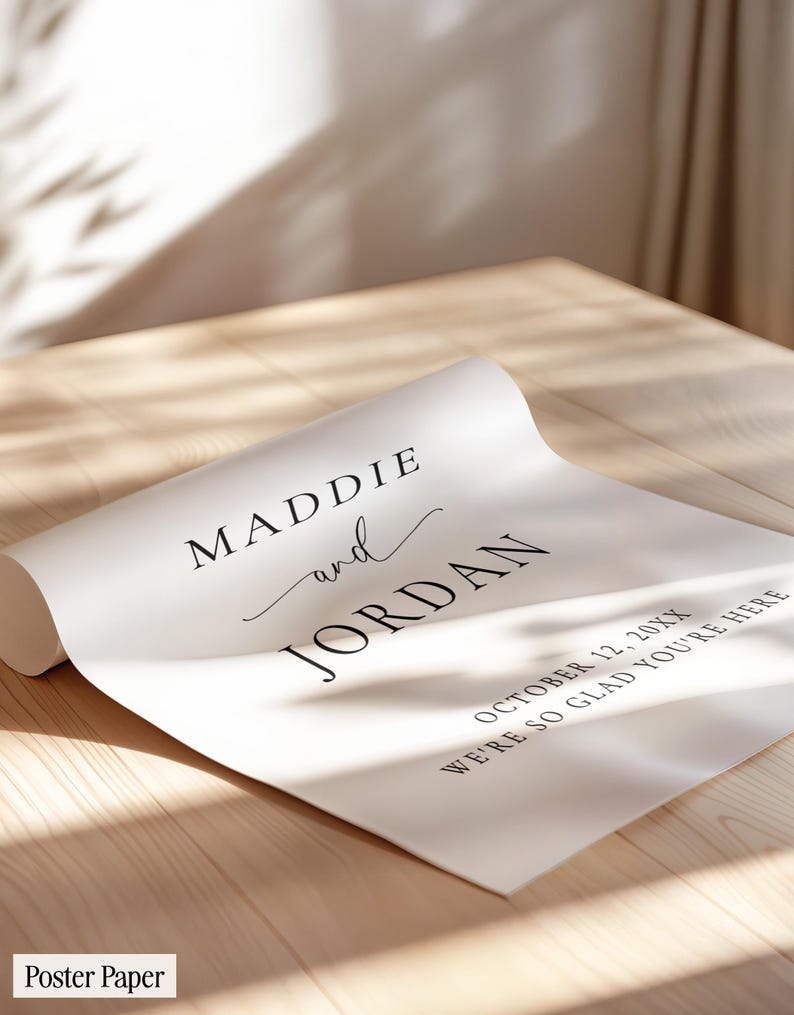 May include: A white poster paper featuring black printed text that reads MADDIE and JORDAN, followed by the date OCTOBER 12, 20XX and the phrase WE'RE SO GLAD YOU'RE HERE. The paper is partially rolled at one end on a light wood surface.
