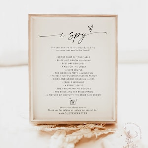 May include: A white framed sign with the text "i spy" and a list of things to find in a wedding photo booth. The sign includes the hashtag #HADLEYEVERAFTER.
