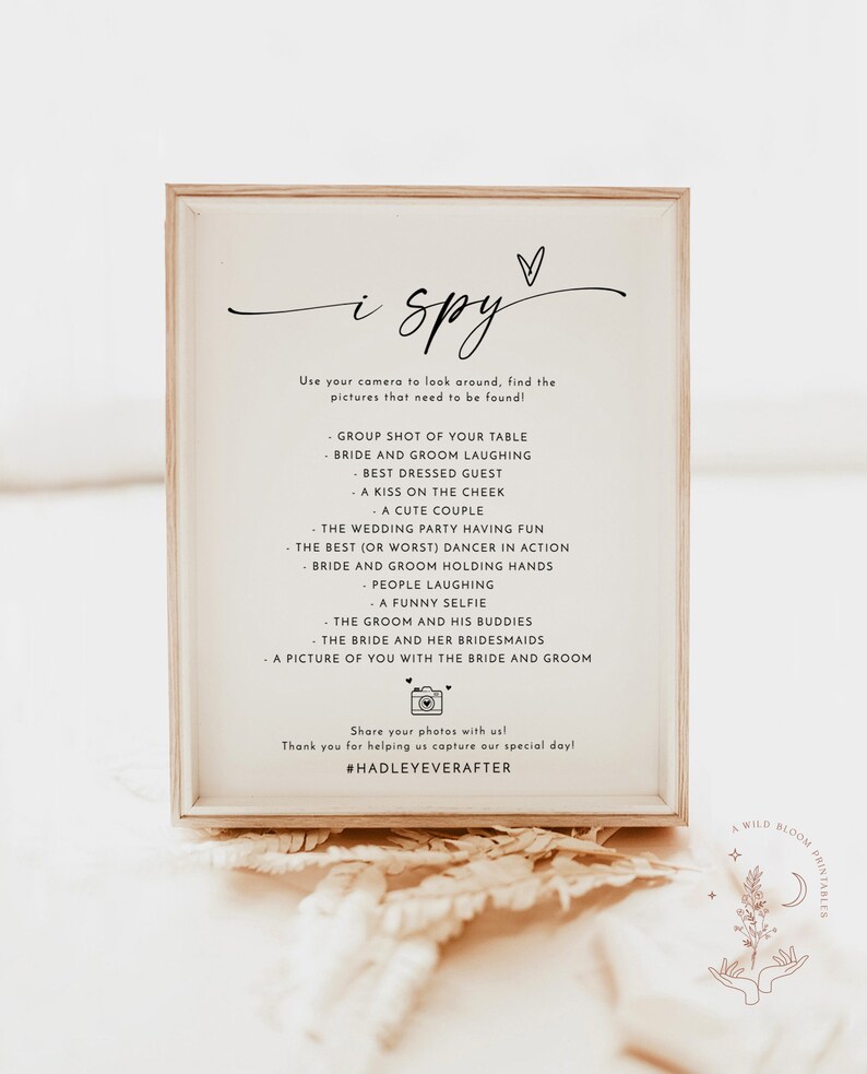 May include: A white framed sign with the text "i spy" and a list of things to find in a wedding photo booth. The sign includes the hashtag #HADLEYEVERAFTER.