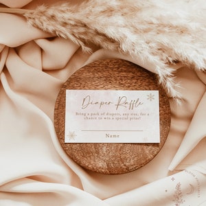 Op de afbeelding: Een baby shower luier verloting kaart met de tekst "Diaper Raffle" in een sierlijke schrift. De kaart is geplaatst op een houten onderzetter en omgeven door zachte, beige stof en gedroogd pampasgras. De kaart bevat ruimte voor een naam.