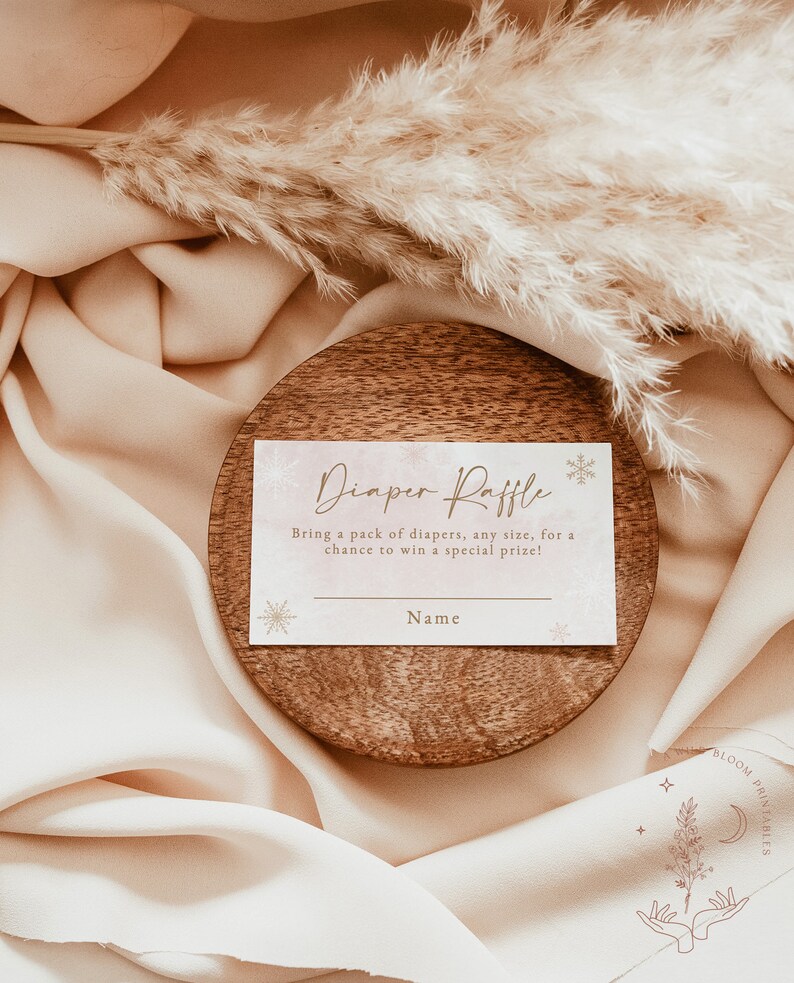 Op de afbeelding: Een baby shower luier verloting kaart met de tekst "Diaper Raffle" in een sierlijke schrift. De kaart is geplaatst op een houten onderzetter en omgeven door zachte, beige stof en gedroogd pampasgras. De kaart bevat ruimte voor een naam.