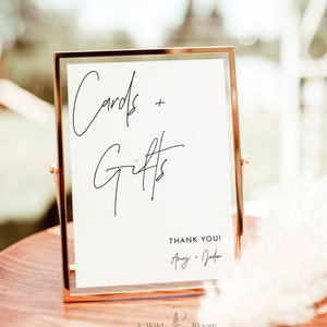 May include: A gold-framed sign with the text "Cards + Gifts" in black cursive writing. Below the text is "Thank You!" followed by the names "Amy + Jordan" in cursive writing. The sign is on a wooden table with a white, fluffy plant in the background.