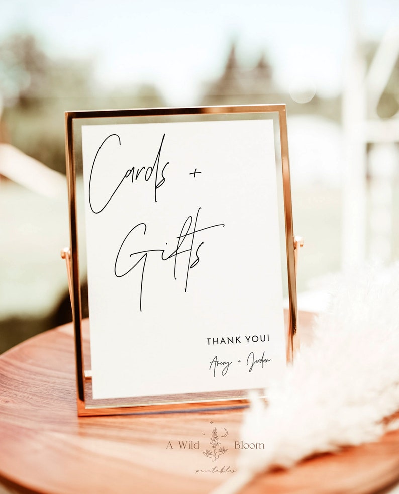 May include: A gold-framed sign with the text "Cards + Gifts" in black cursive writing. Below the text is "Thank You!" followed by the names "Amy + Jordan" in cursive writing. The sign is on a wooden table with a white, fluffy plant in the background.