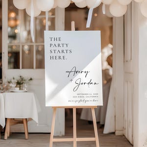 May include: A white rectangular event sign on a light wood easel. The sign features black text reading THE PARTY STARTS HERE, Avery + Jordan, September 24, 20XX, San Diego, California, and we're so glad you're here in a script font.