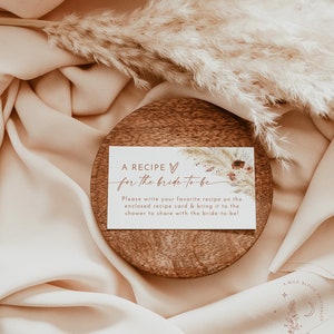 Pu&ograve; includere: Un biglietto bianco con testo marrone che recita "A RECIPE for the bride-to-be". Il biglietto &egrave; su un sottobicchiere di legno con sfondo marrone. Il testo sul biglietto chiede agli ospiti di scrivere la loro ricetta preferita sulla scheda ricetta allegata e di portarla alla doccia per condividerla con la sposa.