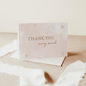 Op de afbeelding: Een gevouwen bedankkaart met de woorden "THANK YOU very much" in gouden letters. De kaart heeft een zachte roze aquarel achtergrond met gouden sneeuwvlok accenten. Een envelop en wit lint zijn ook zichtbaar.