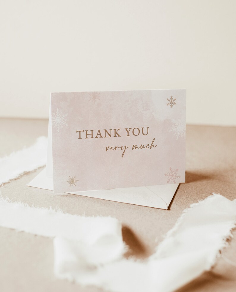 Op de afbeelding: Een gevouwen bedankkaart met de woorden "THANK YOU very much" in gouden letters. De kaart heeft een zachte roze aquarel achtergrond met gouden sneeuwvlok accenten. Een envelop en wit lint zijn ook zichtbaar.