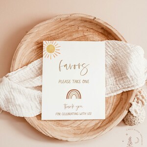 May include: A white card with the word "favors" written in brown cursive script. The card has a sun and rainbow graphic and the text "Please take one", "Thank you", and "For celebrating with us!"