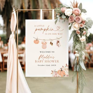 May include: A rectangular 18x24 inch foam board sign for a baby shower. The sign features the text A LITTLE pumpkin IS ON HER WAY, WELCOME TO Maddie's BABY SHOWER, and October 22nd, 20XX. Graphics include a clothesline with a pumpkin, onesie, and shoes.
