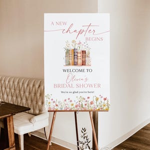 May include: A 24x36 inch foam board sign on a wooden easel. The sign reads "A New Chapter Begins" above "Welcome to Olivia's Bridal Shower." Floral and book illustrations decorate the sign, with the text "We're so glad you're here!"