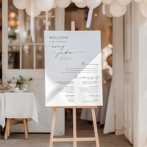 May include: A white rectangular wedding welcome sign on a light wood easel. The sign features black text reading Welcome to the wedding of Avery + Jordan, 09.25.20XX, San Diego, CA, followed by the ceremony schedule and the wedding party list.