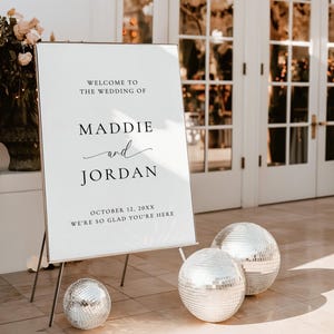 May include: A white wedding welcome sign on a metal stand with black text reading Welcome to the wedding of Maddie and Jordan, October 12, 20XX, We're so glad you're here. Three silver mirrored disco balls are placed on the ground nearby.
