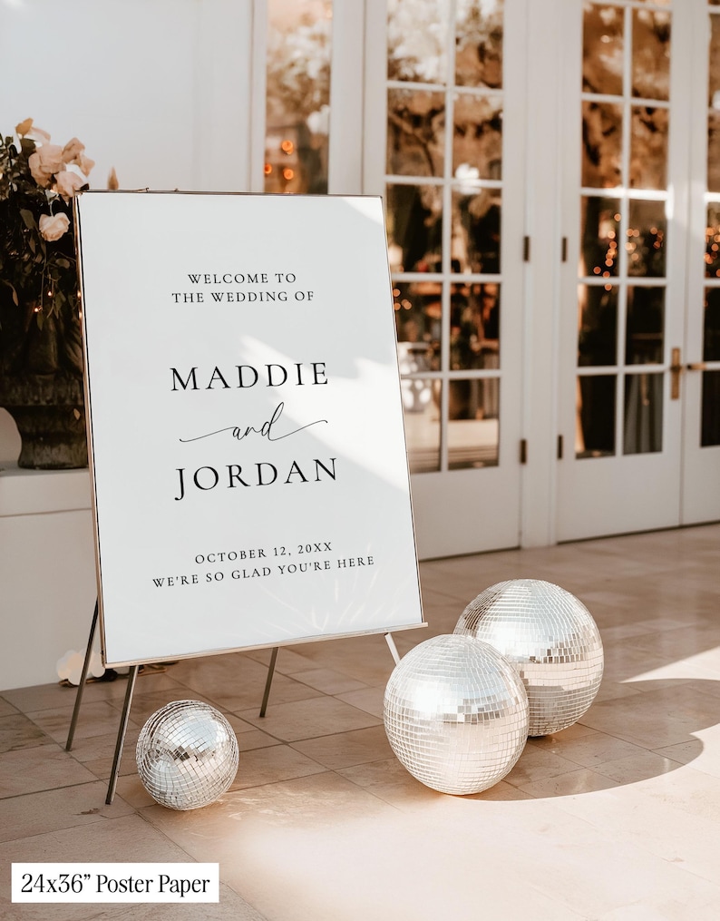 May include: A white wedding welcome sign on a metal stand with black text reading Welcome to the wedding of Maddie and Jordan, October 12, 20XX, We're so glad you're here. Three silver mirrored disco balls are placed on the ground nearby.
