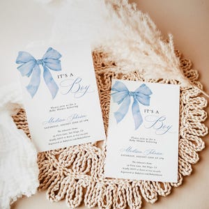 May include: Two blue and white baby shower invitations with a watercolor bow and the text "It's a Boy". The invitations are on a woven placemat.